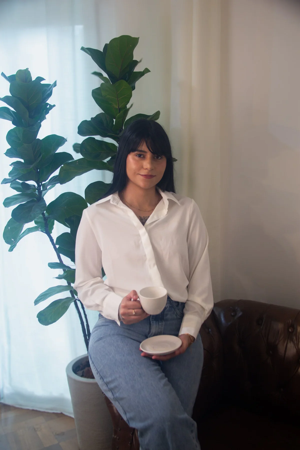 Luany Avela sentada ao lado de uma planta grande, de camisa branca e jeans, segurando xícara e pires